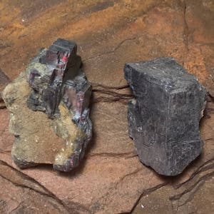May include: Two pieces of black hematite, one with a rough, natural surface and the other with a more polished, crystalline surface. The hematite is a dark, metallic mineral with a silvery sheen.