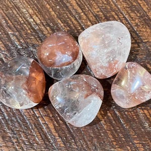 May include: Five tumbled stones with a clear, translucent appearance and reddish-brown inclusions. The stones are smooth and rounded, with varying shapes and sizes. They are arranged on a dark wooden surface.