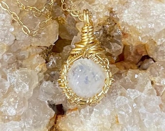 May include: A gold-colored wire-wrapped pendant necklace featuring a round, iridescent moonstone. The moonstone is encased in a woven gold wire setting and hangs from a delicate gold chain. The background is a light-colored, textured surface.