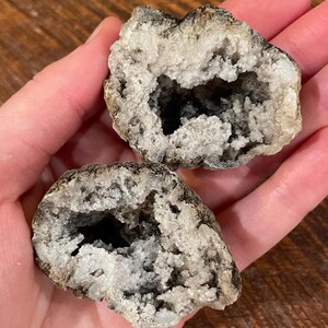 May include: Two halves of a geode, revealing a dark interior cavity lined with sparkling white crystals. The outer shell is a mix of gray and brown, creating a natural, textured appearance. The geode halves are held in a hand.