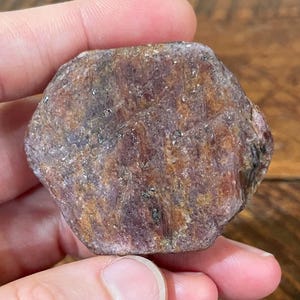 May include: A rough, uncut gemstone with a hexagonal shape. The stone has a mottled appearance with shades of brown, red, and purple. The surface has a slightly rough texture. The gemstone is held in a hand.