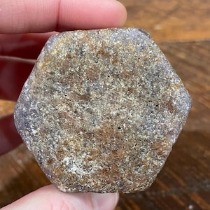 May include: A rough-cut, hexagonal gemstone with a mottled appearance. The stone features a mix of brown, tan, and gray hues, with a slightly textured surface. The overall shape is geometric, and the stone appears to be natural.
