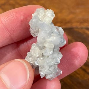 May include: A cluster of pale blue and white crystal formations. The crystals have a rough, textured appearance and are held in a hand. The crystals have a translucent quality, with light passing through them. The overall shape is irregular and organic.