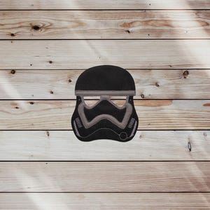May include: A black and gray Stormtrooper helmet design. The helmet features a rounded top and a detailed face mask with gray accents. The design is set against a wooden background.