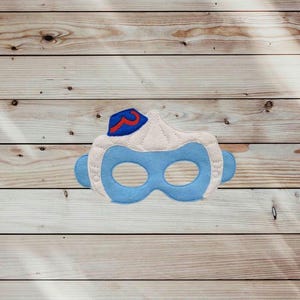 May include: A felt mask featuring a light blue base with two eye openings, topped with a cream-colored section and a small blue hat detail accented with a red curved line. The mask has visible white stitching along the edges of the fabric layers.