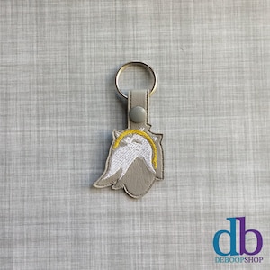 May include: A gray and white embroidered keychain with a yellow accent. The keychain features a stylized design and a silver key ring. The keychain is attached to a gray leather strap with a snap closure.