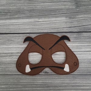 May include: A brown felt mask with black detailing, including eye holes and a downturned mouth. The mask has two white tusks and is designed to resemble a cartoon character. The mask is likely for costume or play.