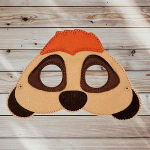 May include: A felt mask shaped like a meerkat face, featuring a tan base, a bright orange patch on the forehead, dark brown eye patches surrounding the circular eye holes, a dark brown nose, and dark brown accents on the outer edges.