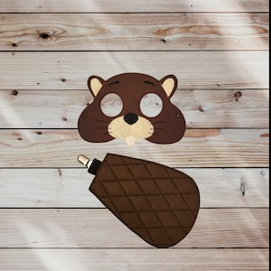 May include: A brown felt beaver mask with a beige muzzle and a small white tooth. Below the mask is a brown felt beaver tail with a diamond pattern and a silver clip. The items are on a wooden background.