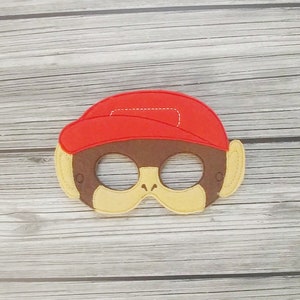 May include: A felt monkey mask with a red hat, brown face, and tan accents. The mask has eye holes and is designed to be worn. The mask is likely for a child's costume or play.