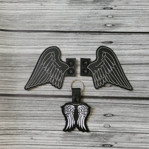 May include: Black embroidered angel wing patches and a keychain with a matching wing design. The wings are outlined with white stitching. The patches have a button-like design for attachment. The keychain has a black leather strap and silver-colored ring.