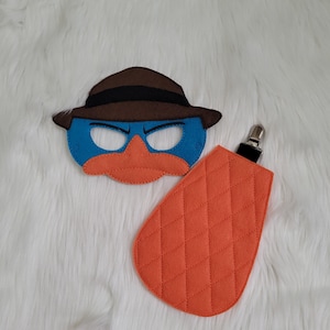 May include: A blue and orange felt mask with a brown hat, and an orange quilted pacifier clip. The mask is in the shape of a platypus. The pacifier clip has a metal clip.