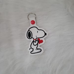 May include: White Snoopy keychain holding a red heart. The keychain has a red button and a silver metal ring. The Snoopy design is outlined in black and has black ears. The background is a soft, white, fluffy surface.