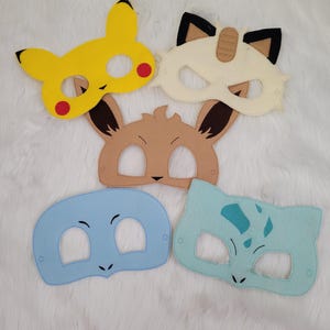 May include: Five colorful felt masks in the shape of popular cartoon characters. The masks include a yellow Pikachu, a cream-colored cat, a brown Eevee, a blue mask, and a light blue mask with dark blue spots.