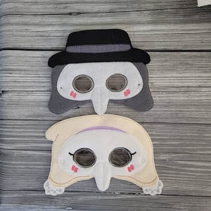 May include: Two felt masks, one grey with a black hat and the other cream coloured. Both masks have a long nose and eye holes with pink accents.