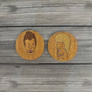 May include: Two round cork coasters featuring cartoon character portraits. One coaster has a brown-haired character with an open mouth, and the other has a blonde-haired character with a smirk. The coasters are made of natural cork material.