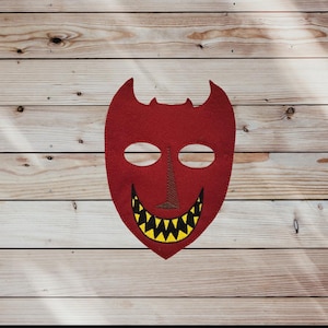 May include: A red felt mask with a devilish design. The mask features two eye holes, a stitched brown nose, and a wide, toothy grin with yellow teeth. The mask has two curved horns at the top.