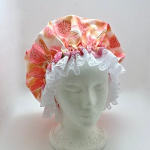 May include: A pink and white floral shower cap with a white lace trim. The cap is on a white mannequin head.