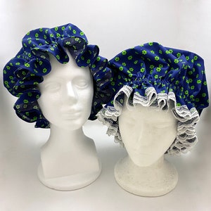 May include: Two blue shower caps with a green floral pattern are displayed on white mannequin heads. One cap has a ruffled edge, while the other has a white lace trim. The caps are designed to protect hair from water.