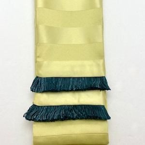 May include: A yellow and green striped fabric sash with a dark green fringe trim. The sash is folded in thirds and the fringe is visible on each fold.