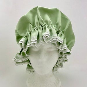 May include: A light green bonnet with a ruffled design and white lace trim. The bonnet is displayed on a white mannequin head. The fabric has a textured appearance, and the overall style is vintage.