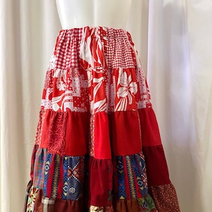 May include: A red, white, and burgundy patchwork skirt with multiple tiers. The skirt is made from a variety of fabrics, including gingham, floral, and geometric patterns.