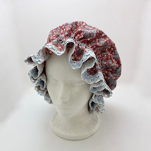 May include: A floral bonnet with a ruffled brim. The bonnet features a red fabric with a blue floral pattern. The brim is trimmed with blue lace. The bonnet is displayed on a white mannequin head.