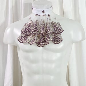 May include: A white ruffled ascot collar with a decorative design. The collar is displayed on a white mannequin. The ascot features a white collar with purple and gold accents and a cascading, layered ruffle design.
