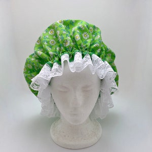 May include: Green shower cap with a white lace trim and a daisy print. The cap is on a white mannequin head.