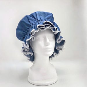 May include: A blue denim bonnet with a ruffled brim and white lace trim. The bonnet is displayed on a white mannequin head against a white background. The hat has a gathered crown and a wide, decorative brim.