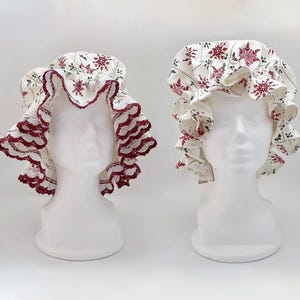 May include: Two white fabric bonnets with red snowflake patterns and ruffled edges are displayed on white mannequin heads. One bonnet has a red lace trim, while the other has a matching red snowflake pattern on the trim. The bonnets are likely for hair protection or fashion.