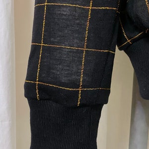 May include: Black long-sleeved shirt with a gold grid pattern. The shirt has a ribbed cuff.