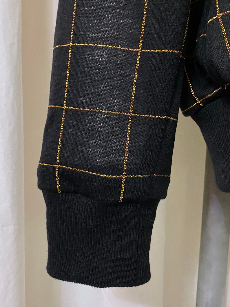 May include: Black long-sleeved shirt with a gold grid pattern. The shirt has a ribbed cuff.