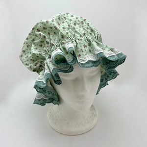 May include: A light green bonnet with a floral pattern of small red flowers and green leaves. The bonnet features a ruffled brim with white lace trim and a teal-colored edge. The bonnet is displayed on a white mannequin head.