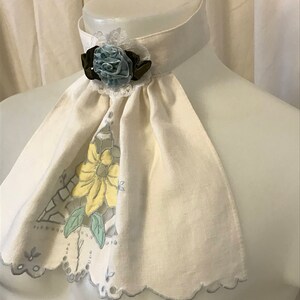 Puede incluir: Un cuello de lino blanco con borde festoneado y dise&ntilde;o floral. Una flor azul y verde con un acento marr&oacute;n est&aacute; unida al cuello.