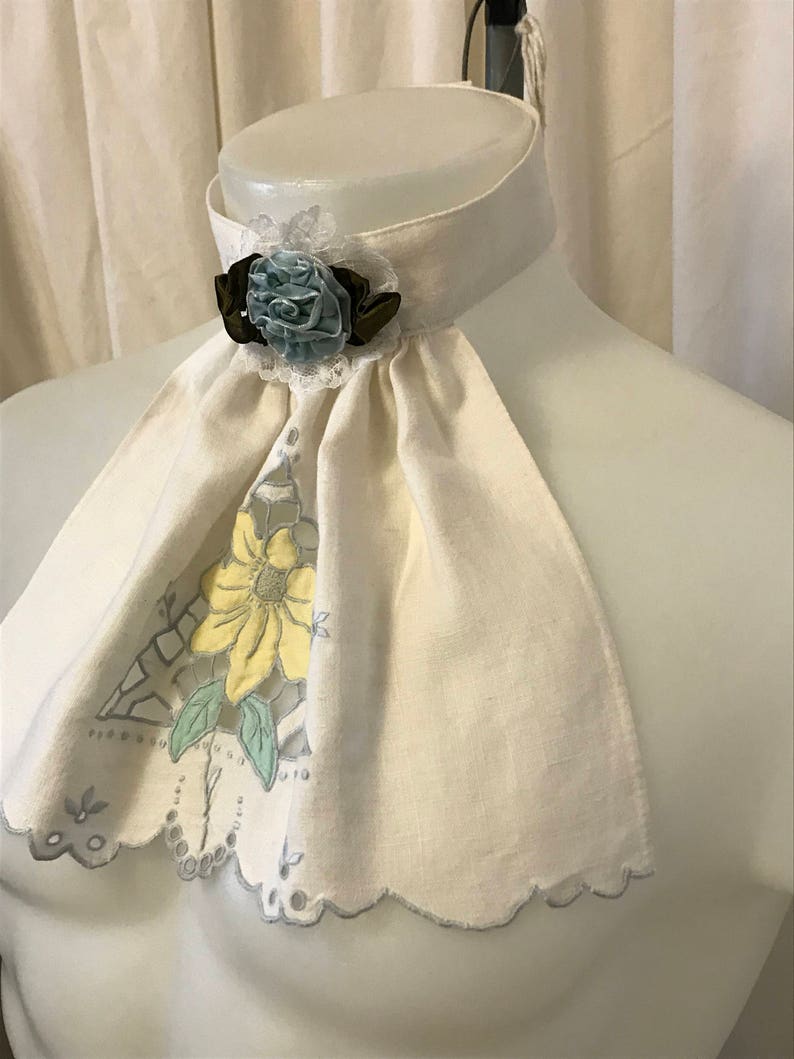 Puede incluir: Un cuello de lino blanco con borde festoneado y dise&ntilde;o floral. Una flor azul y verde con un acento marr&oacute;n est&aacute; unida al cuello.
