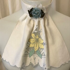 Puede incluir: Un cuello de lino blanco con borde festoneado y un dise&ntilde;o floral amarillo. Una flor de tela azul y marr&oacute;n est&aacute; unida al cuello.