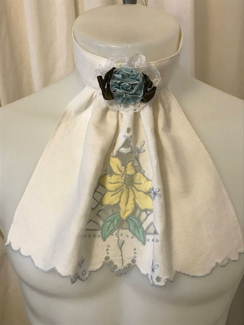 Puede incluir: Un cuello de lino blanco con borde festoneado y un dise&ntilde;o floral amarillo. Una flor de tela azul y marr&oacute;n est&aacute; unida al cuello.