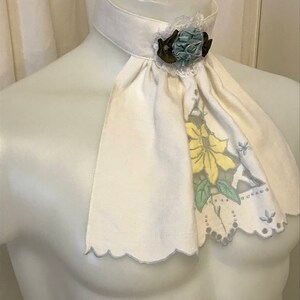 Puede incluir: Una corbata de lino blanco con un borde festoneado y un dise&ntilde;o floral. La corbata tiene un broche de flor de tela azul y marr&oacute;n.
