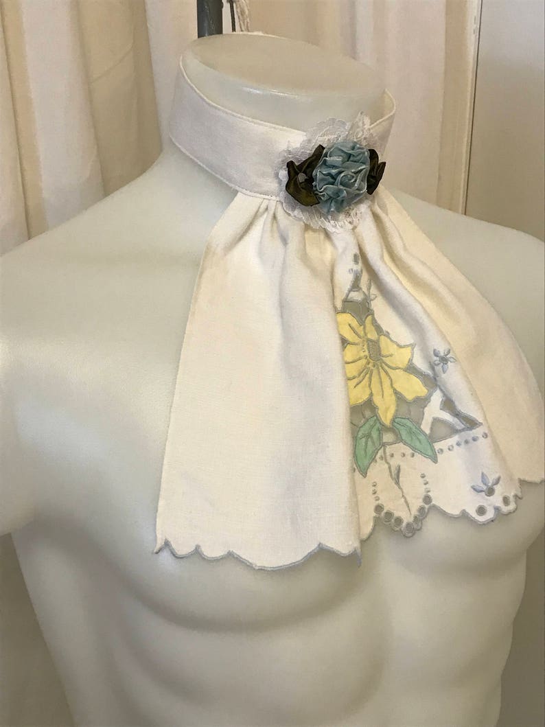 Puede incluir: Una corbata de lino blanco con un borde festoneado y un dise&ntilde;o floral. La corbata tiene un broche de flor de tela azul y marr&oacute;n.
