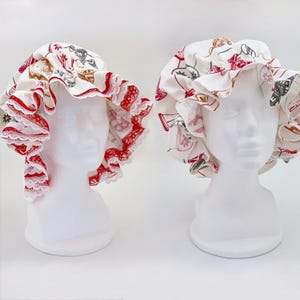 May include: Two white shower caps with a red and white floral pattern and red lace trim. The caps are displayed on white mannequin heads against a white background. The caps have a ruffled edge.