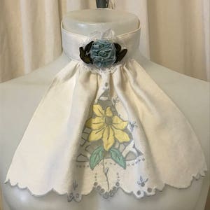 Puede incluir: Un cuello de lino blanco con borde festoneado y un dise&ntilde;o floral azul y amarillo. El cuello tiene una flor de sat&eacute;n azul con un centro marr&oacute;n.