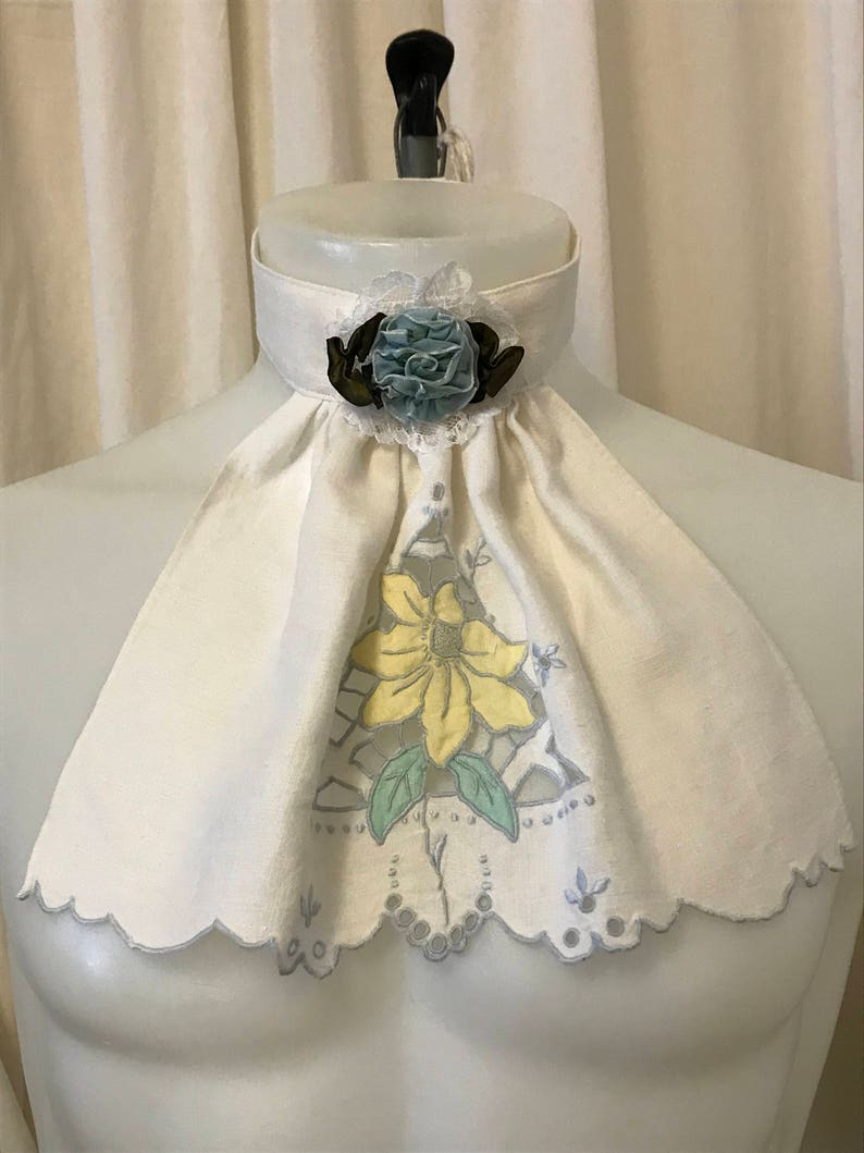 Puede incluir: Un cuello de lino blanco con borde festoneado y un dise&ntilde;o floral azul y amarillo. El cuello tiene una flor de sat&eacute;n azul con un centro marr&oacute;n.