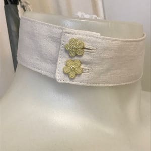 Puede incluir: Un cuello de lino blanco con dos botones dorados en forma de flor.