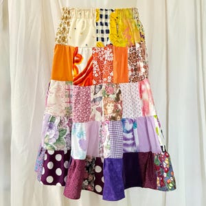May include: A tiered midi skirt featuring a patchwork design of various fabrics in shades of orange, yellow, purple, pink, and white. The skirt includes an elastic waistband and displays diverse patterns such as floral prints, polka dots, gingham, and illustrations of butterflies and faces.