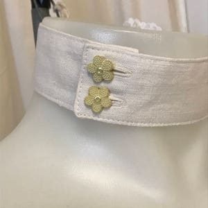 Puede incluir: Un cuello de lino blanco con dos botones dorados en forma de flor.