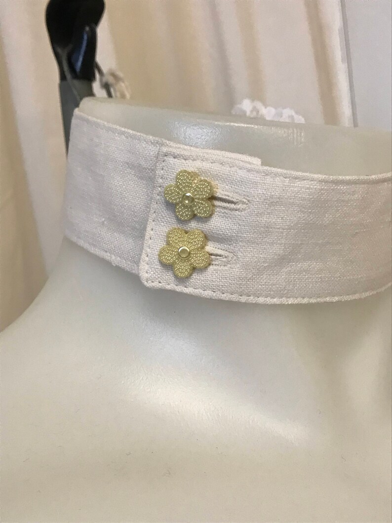 Puede incluir: Un cuello de lino blanco con dos botones dorados en forma de flor.