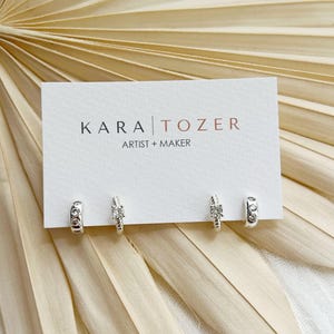 May include: Two pairs of small silver hoop earrings, each featuring a single clear round stone, are attached to a white rectangular display card. The card displays the black and copper text KARA TOZER ARTIST + MAKER in the center.