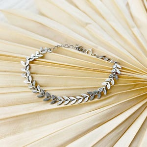 May include: A silver-colored bracelet featuring a delicate leaf design. The bracelet is composed of small, detailed leaves linked together, creating a continuous pattern. The clasp is visible, and the bracelet is displayed on a neutral, textured background.
