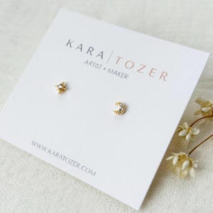 May include: A pair of gold-colored stud earrings, one shaped like a star and the other a crescent moon, displayed on a white card. The star earring has a small, clear stone in the center. The card has the text "KARA TOZER ARTIST + MAKER" and a website address.